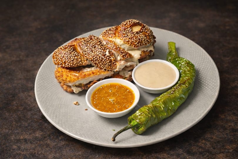 Chicken schnitzel sandwich on a sesame bun with two dipping sauces and a roasted green pepper.