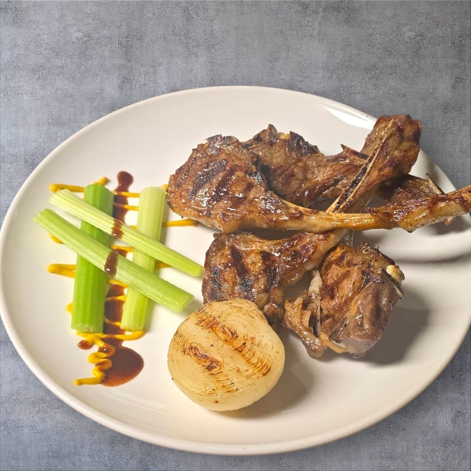 Grilled lamb chops with celery sticks, a grilled onion, and sauces on a white plate.