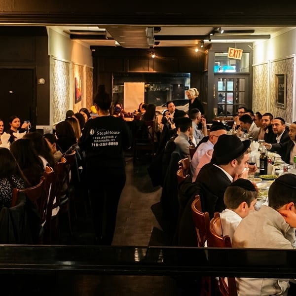 A busy restaurant dining room with many patrons seated at long tables and a server walking through.